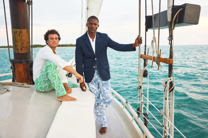 Two men on a sailboat, one in green and white patterned pants and a white shirt, the other in blue patterned pants and a blue blazer.